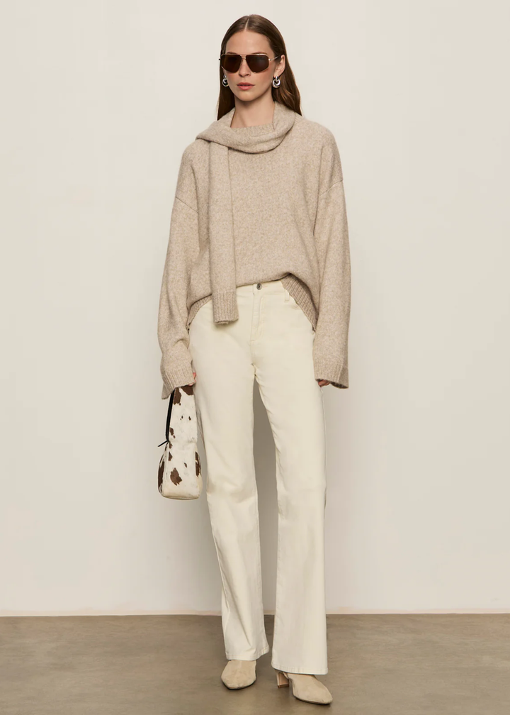 Woman wearing a tan sweater with a tan scarf and cream jeans holding a purse