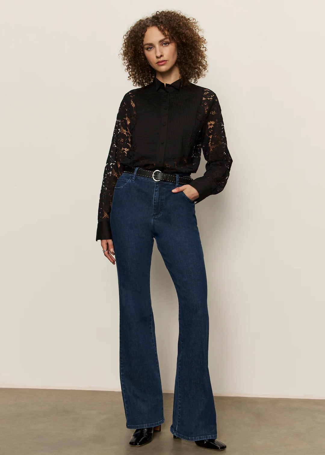 Woman with hand in pocket wearing a black button down shirt with lace sleeves and back with jeans against a white background