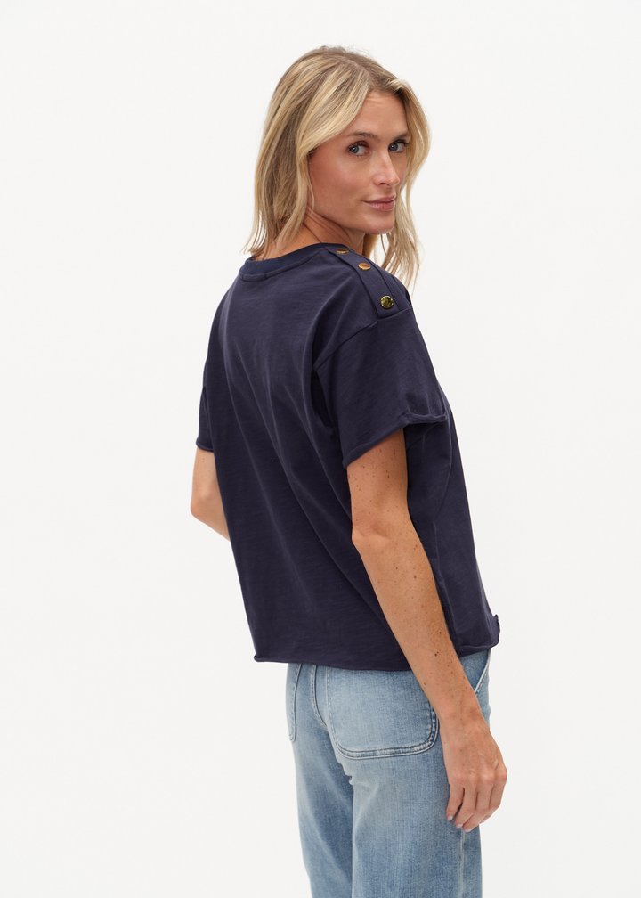 Woman looking over shoulder wearing a navy tee shirt with buttons on the shoulders