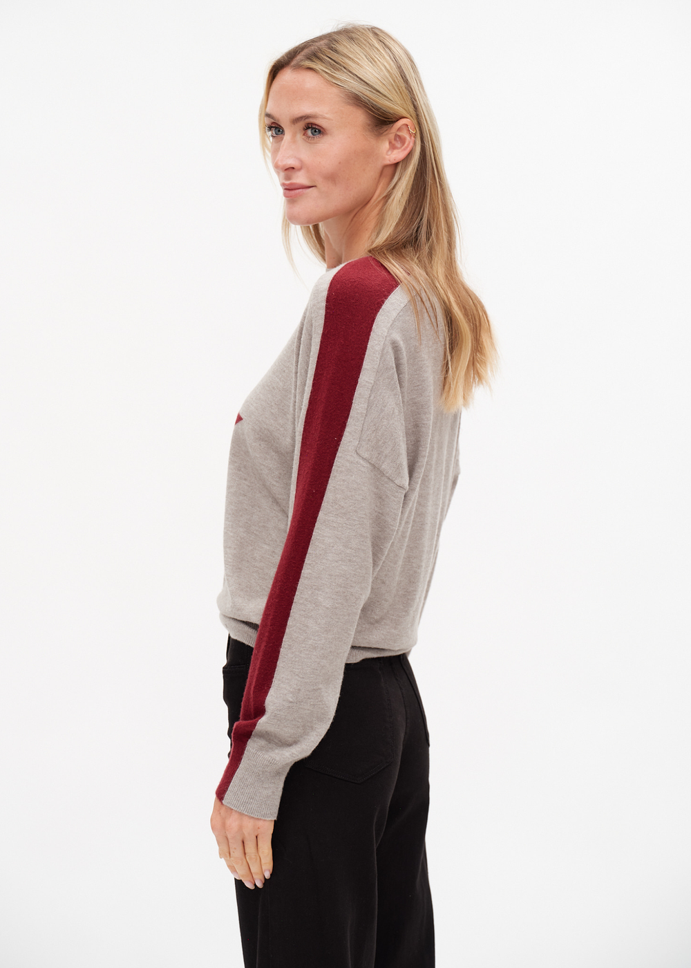 Woman turned to the side wearing a grey sweater with a red star and sleeve stripes against a white background