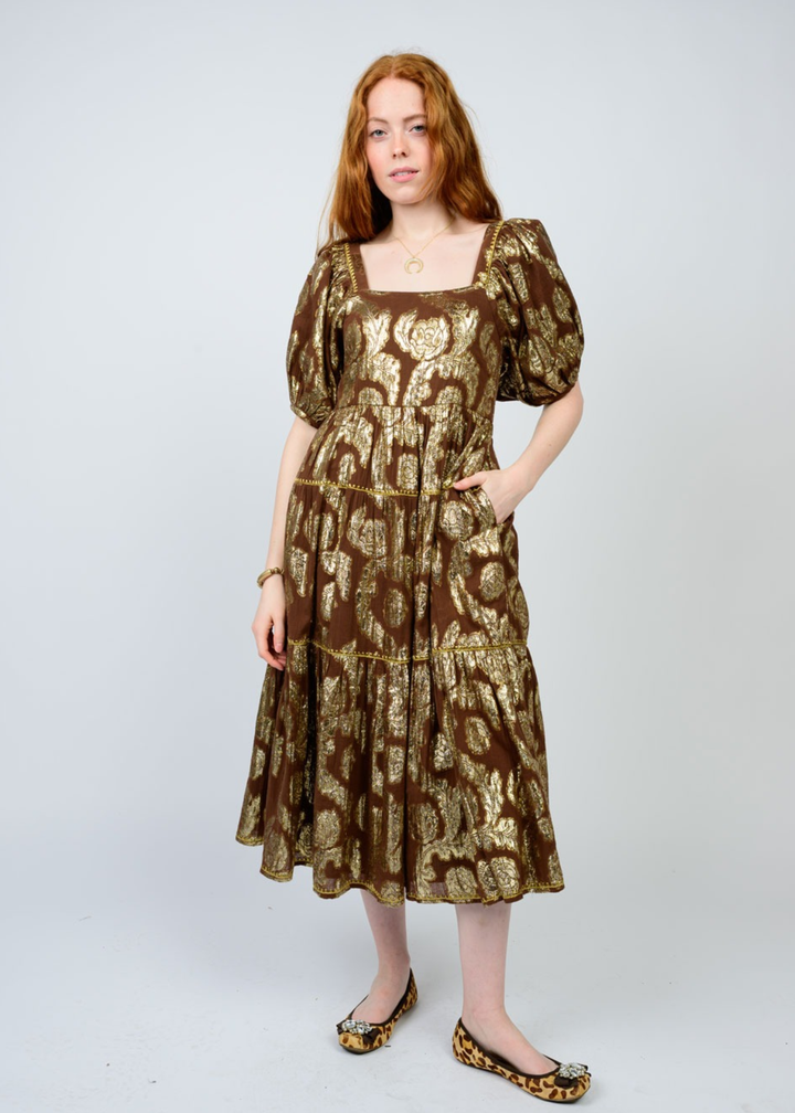 Woman with hand in pocket wearing a brown midi dress with a gold floral pattern