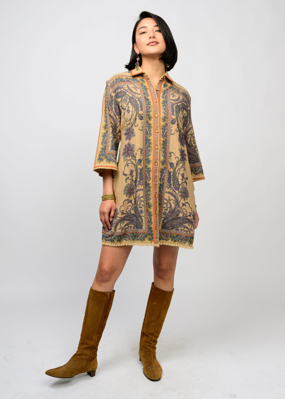 Woman wearing a camel and floral button down dress with 3/4 length sleeves and tall brown boots standing in front of a white background