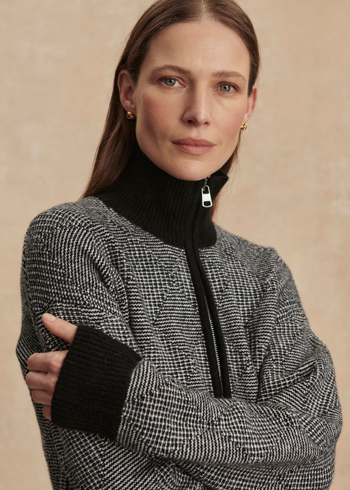 Woman with arms crossed wearing a black knit top with black pants against a tan background