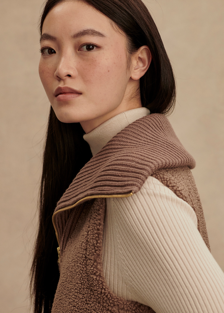 Close up of a woman turned to the side wearing a brown vest over a cream turtleneck in front of a tan background