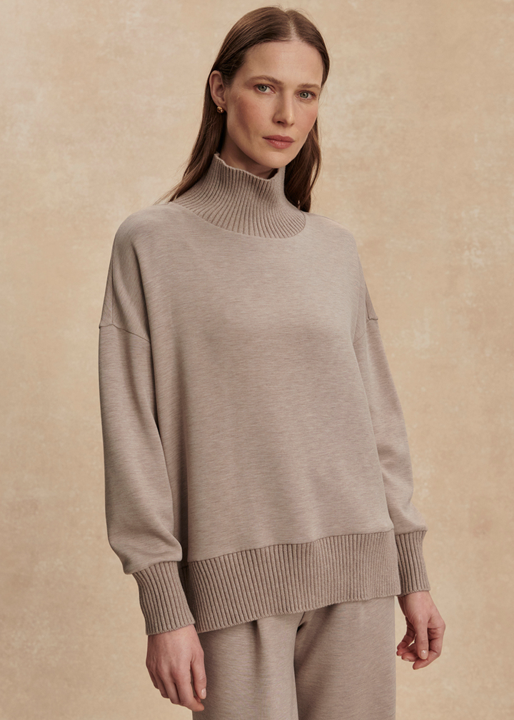 Woman wearing a taupe high neck sweatshirt with taupe pants in front of a tan background