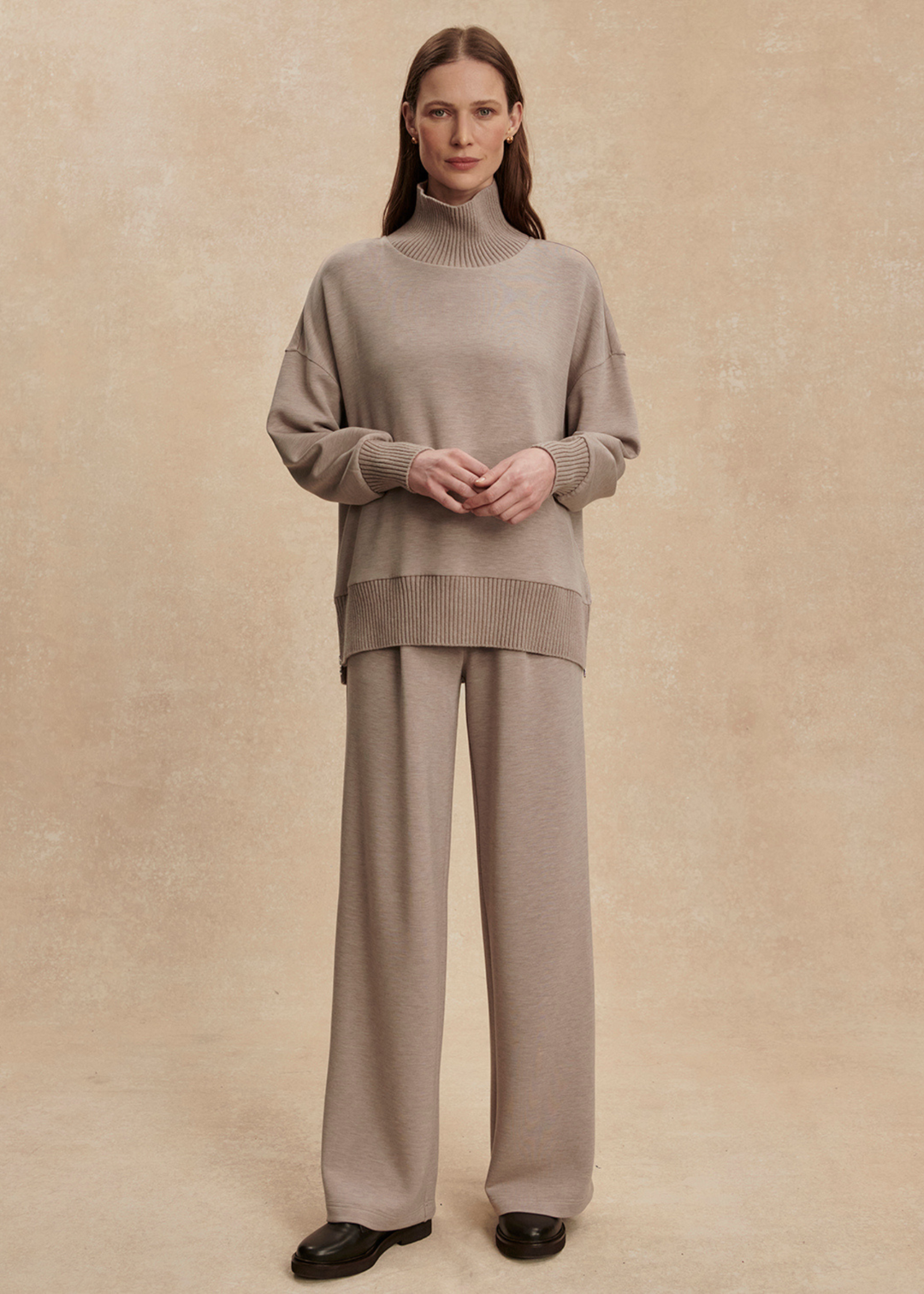 Woman wearing a taupe high neck sweatshirt with taupe pants in front of a tan background