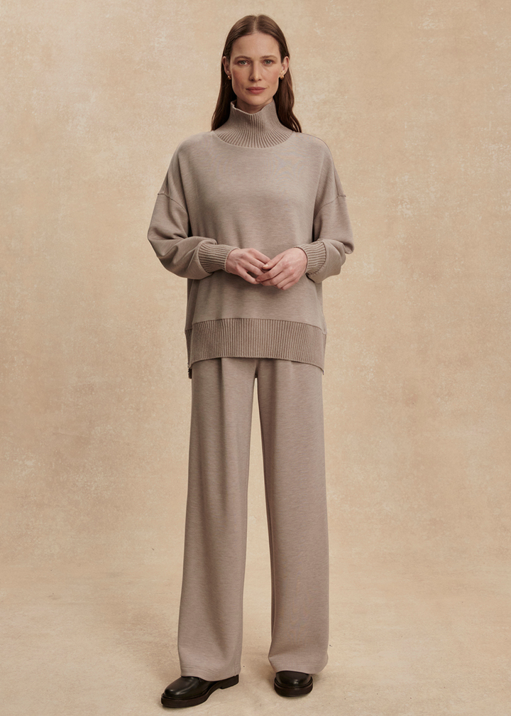 Woman wearing a taupe high neck sweatshirt with taupe pants in front of a tan background