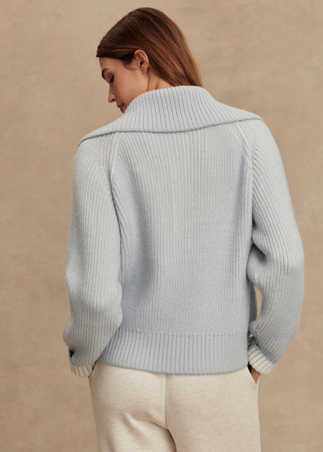 Woman turned around wearing a light blue ribbed sweater with white pants in front of a tan background