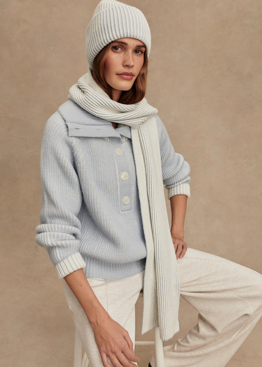 Woman sitting on a white stool wearing a light blue ribbed sweater with white pants and a white and blue ribbed beanie and scarf in front of a tan background