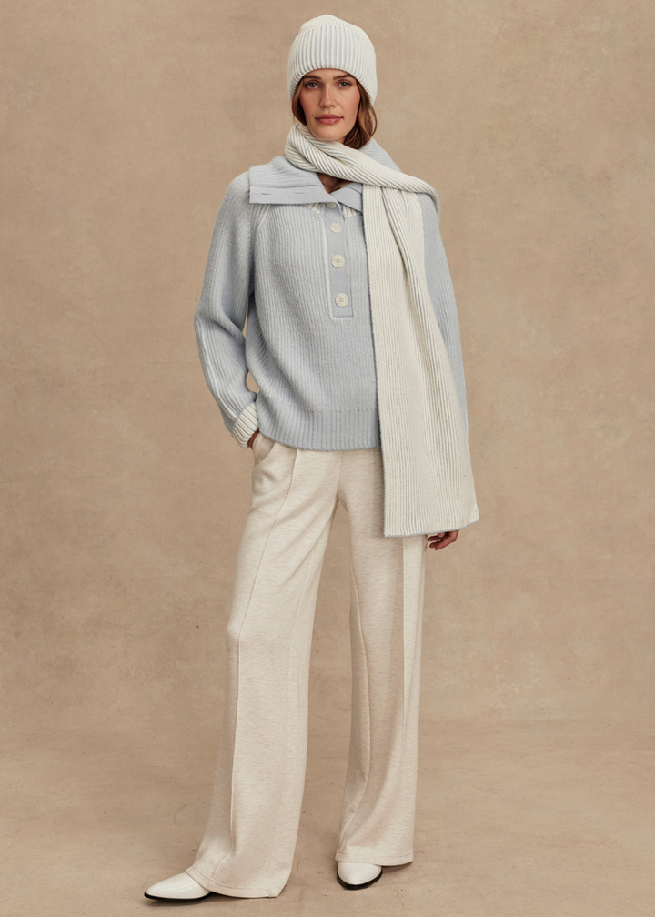 Woman wearing a light blue ribbed sweater with white pants and a white and blue ribbed beanie and scarf in front of a tan background