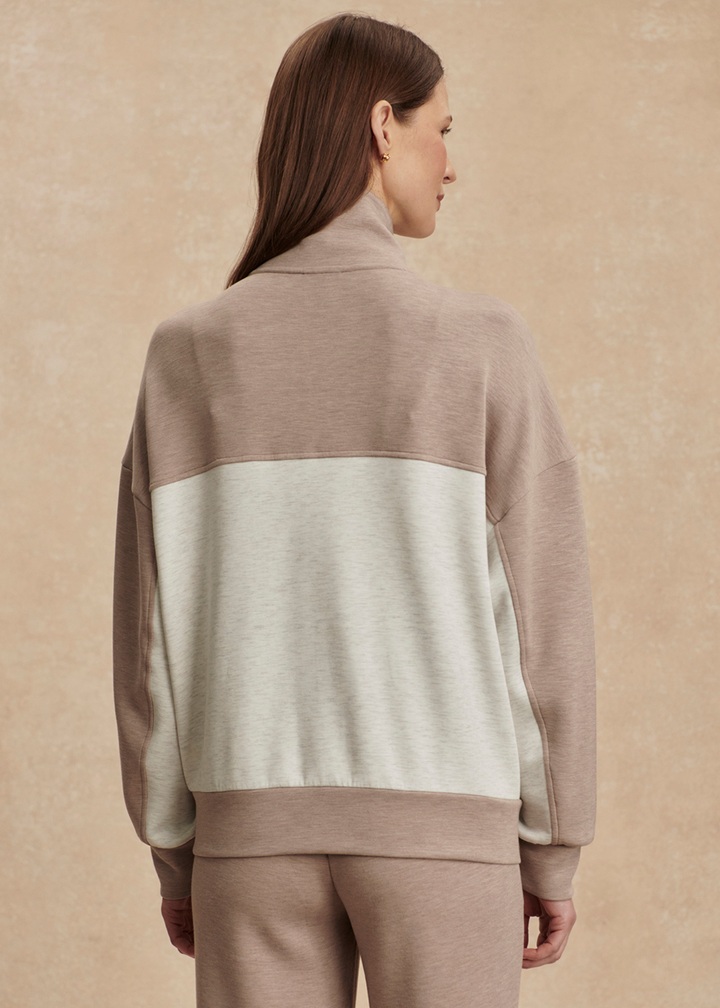 Woman turned around wearing a taupe and white zip up sweatshirt with taupe pants against a tan background