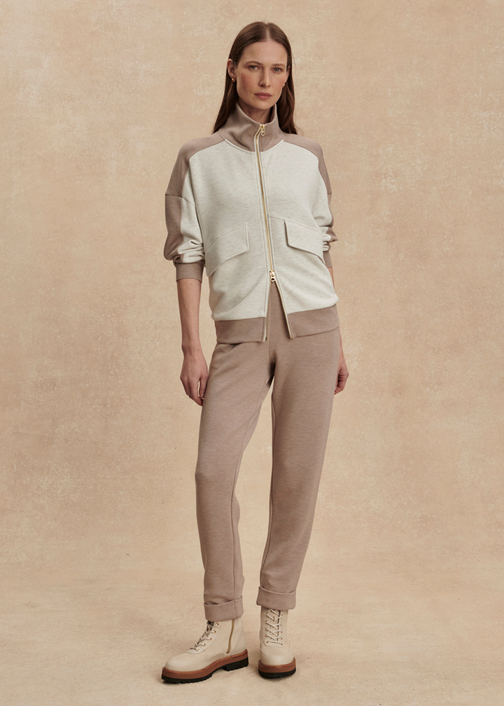Woman wearing a taupe and white zip up sweatshirt with taupe pants against a tan background