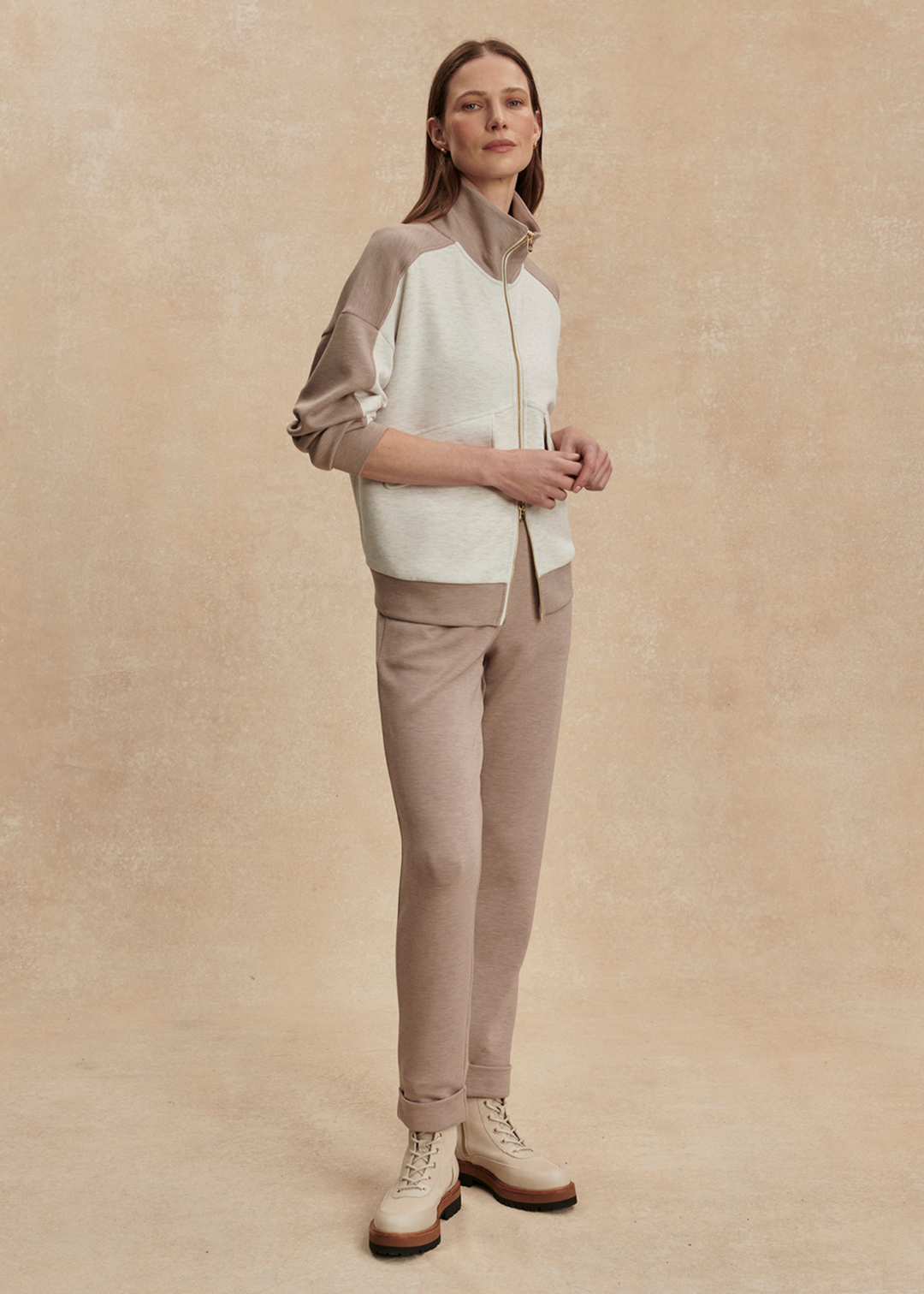 Woman wearing a taupe and white zip up sweatshirt with taupe pants against a tan background