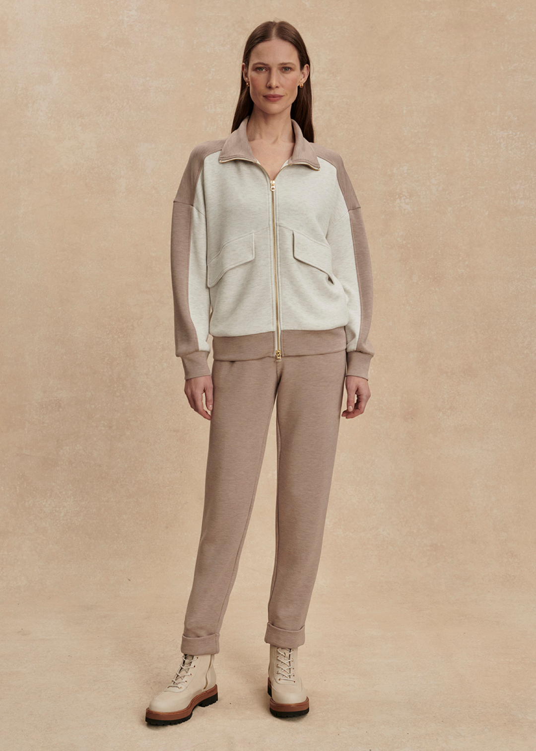 Woman wearing a taupe and white zip up sweatshirt with taupe pants against a tan background