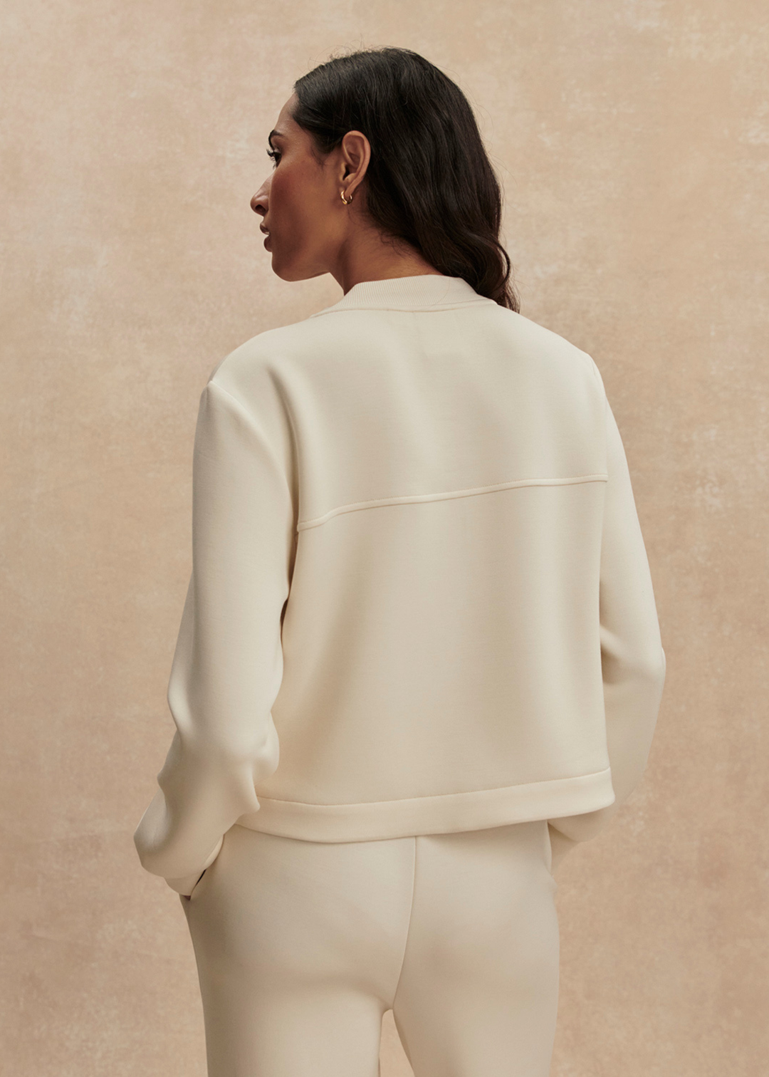 Woman turned around wearing a cream button down sweater with matching pants against a tan background