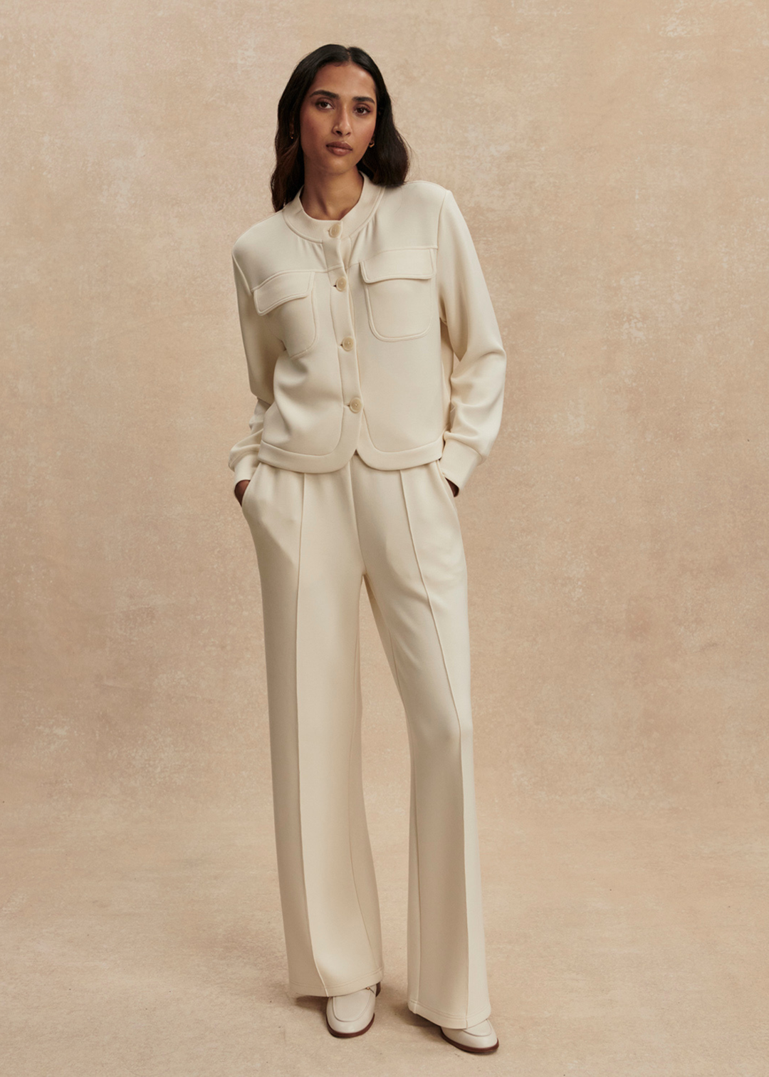 Woman wearing a cream button down sweater with matching pants against a tan background