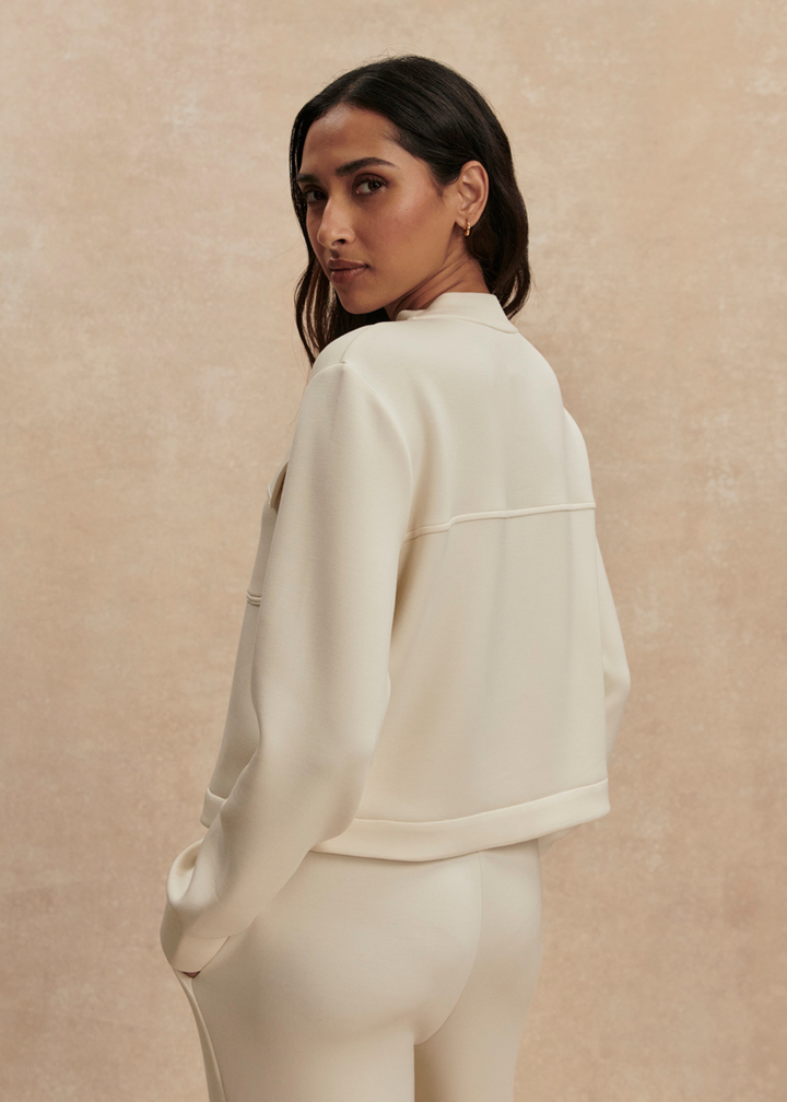 Woman looking over shoulder wearing a cream button down sweater with matching pants against a tan background