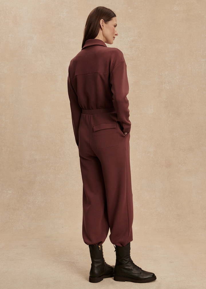 Woman turned to the side wearing a brown jumpsuit with black boots in front of a tan background