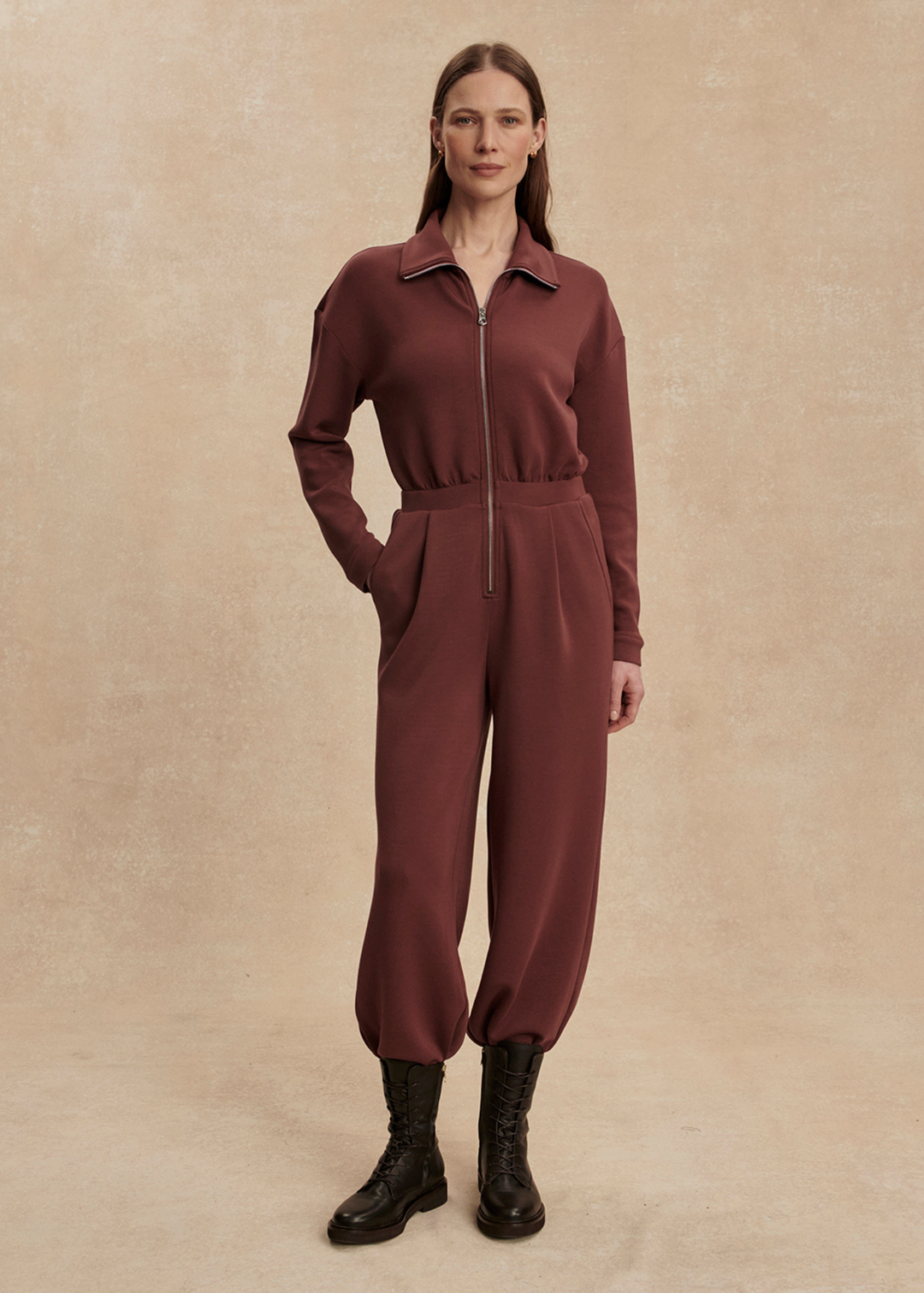 Woman with hand in pocket wearing a brown jumpsuit with black boots in front of a tan background