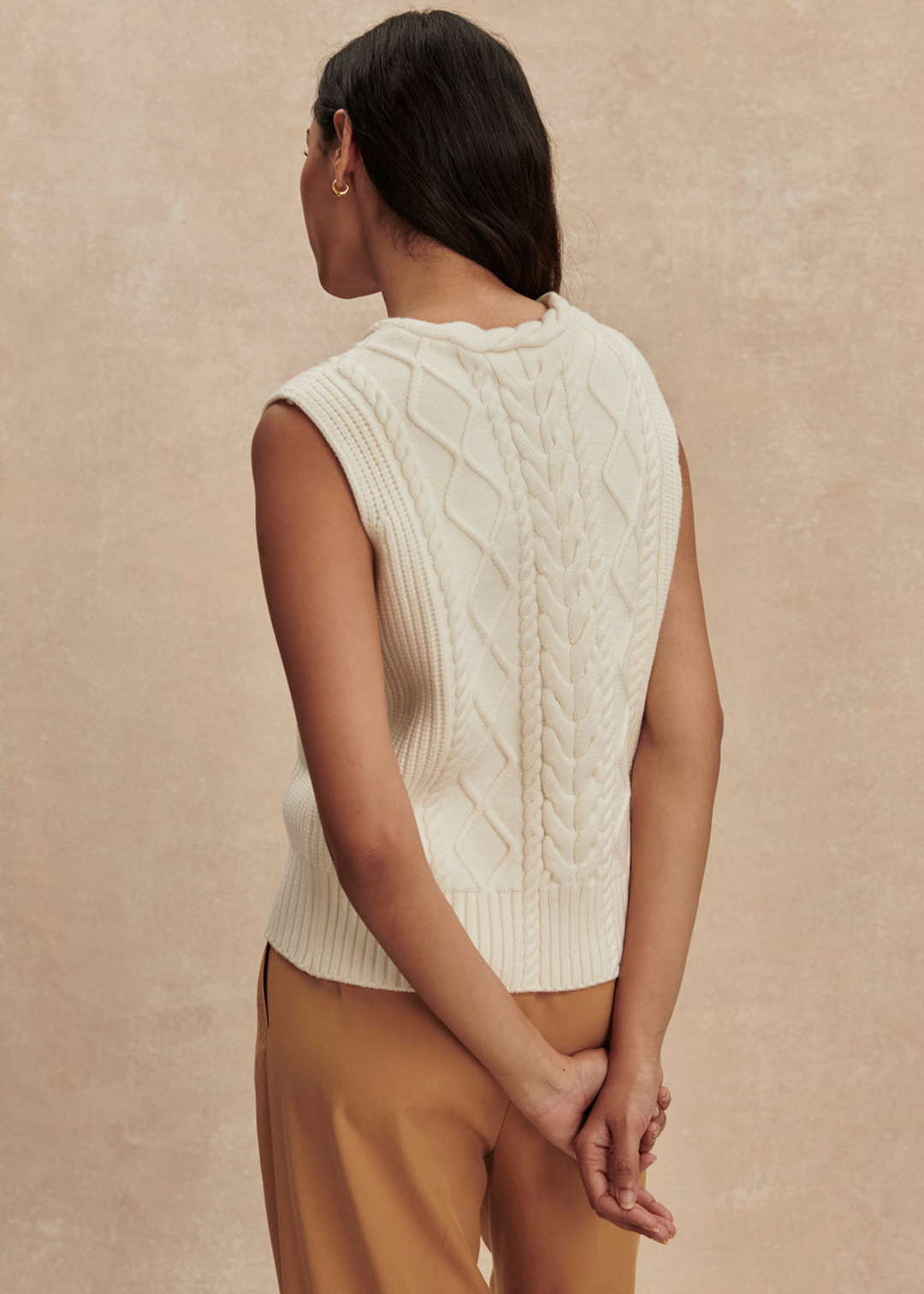 Woman turned around wearing a cream cable knit sweater vest with brown pants