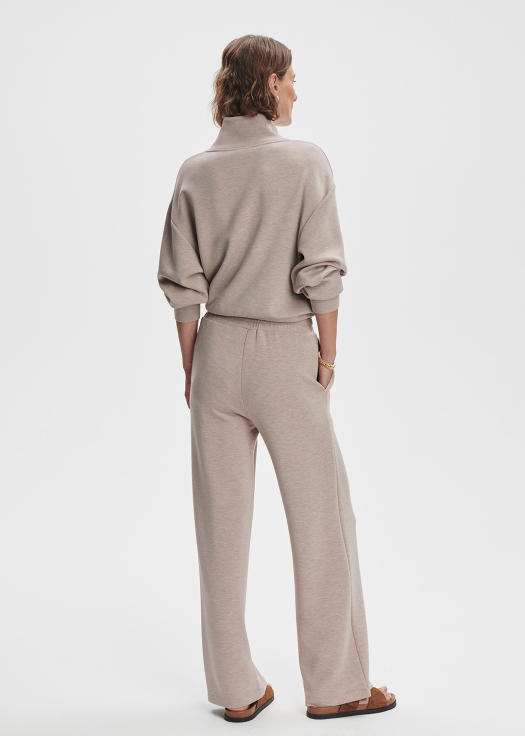 Woman turned around wearing taupe wide leg pants with a taupe half zip top against a white background