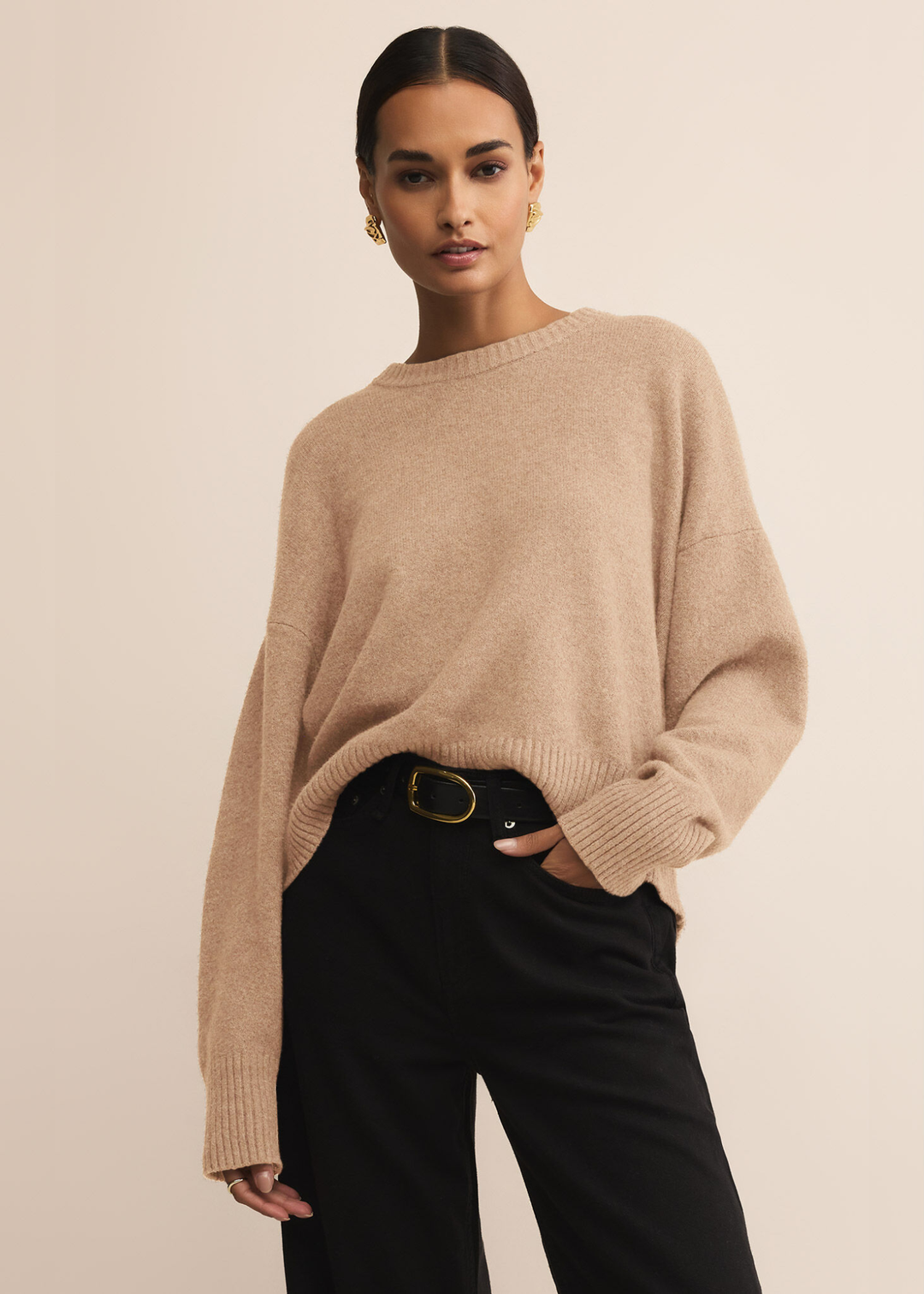 Woman wearing a tan sweater with dark jeans standing in front of a white background