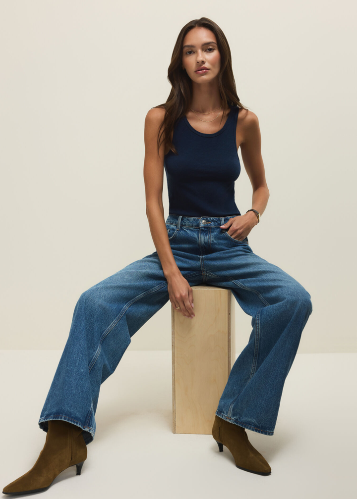 Woman sitting wearing a navy tank top with jeans against a white background