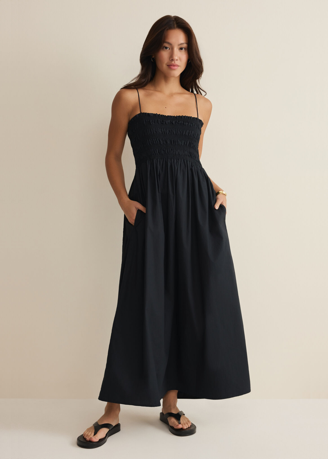 Woman with hands in pockets wearing a black spaghetti strap midi dress against a white background