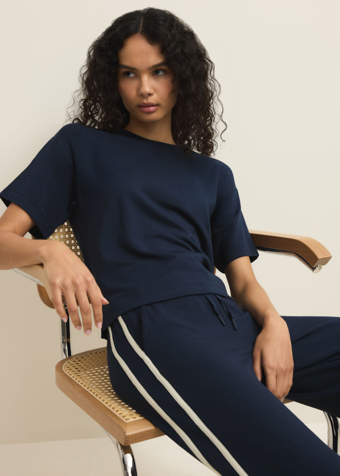 Woman sitting wearing navy sweatpants with white side stripes and a navy tee against a white background