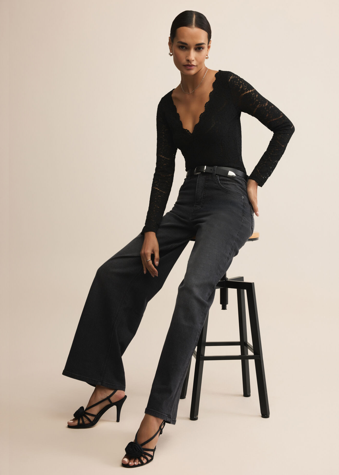 Woman sitting on a stool wearing a black lace bodysuit with scalloped trim and black jeans against a tan background