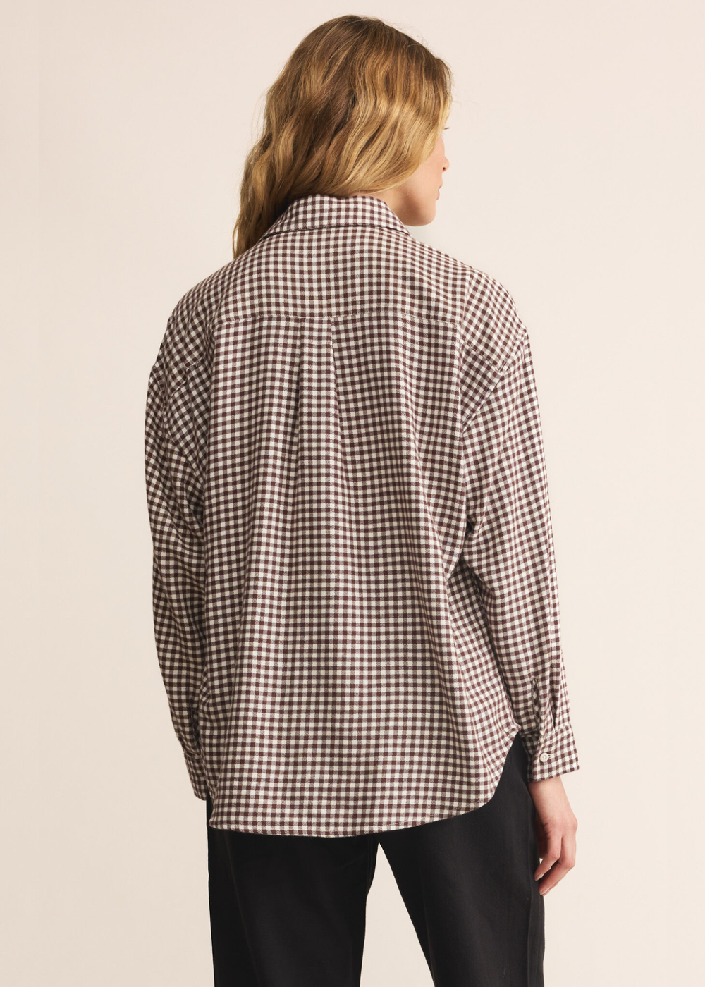 Woman turned around wearing a brown and white plaid button up with dark jeans standing in front of a white background