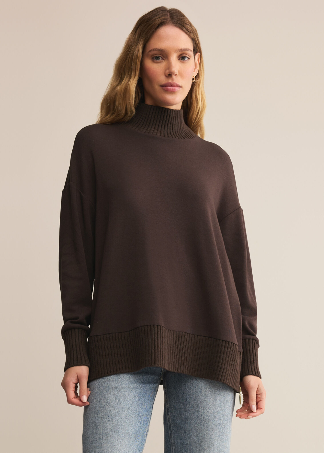 Woman wearing a brown sweater with a mock neckline and jeans standing in front of a tan background
