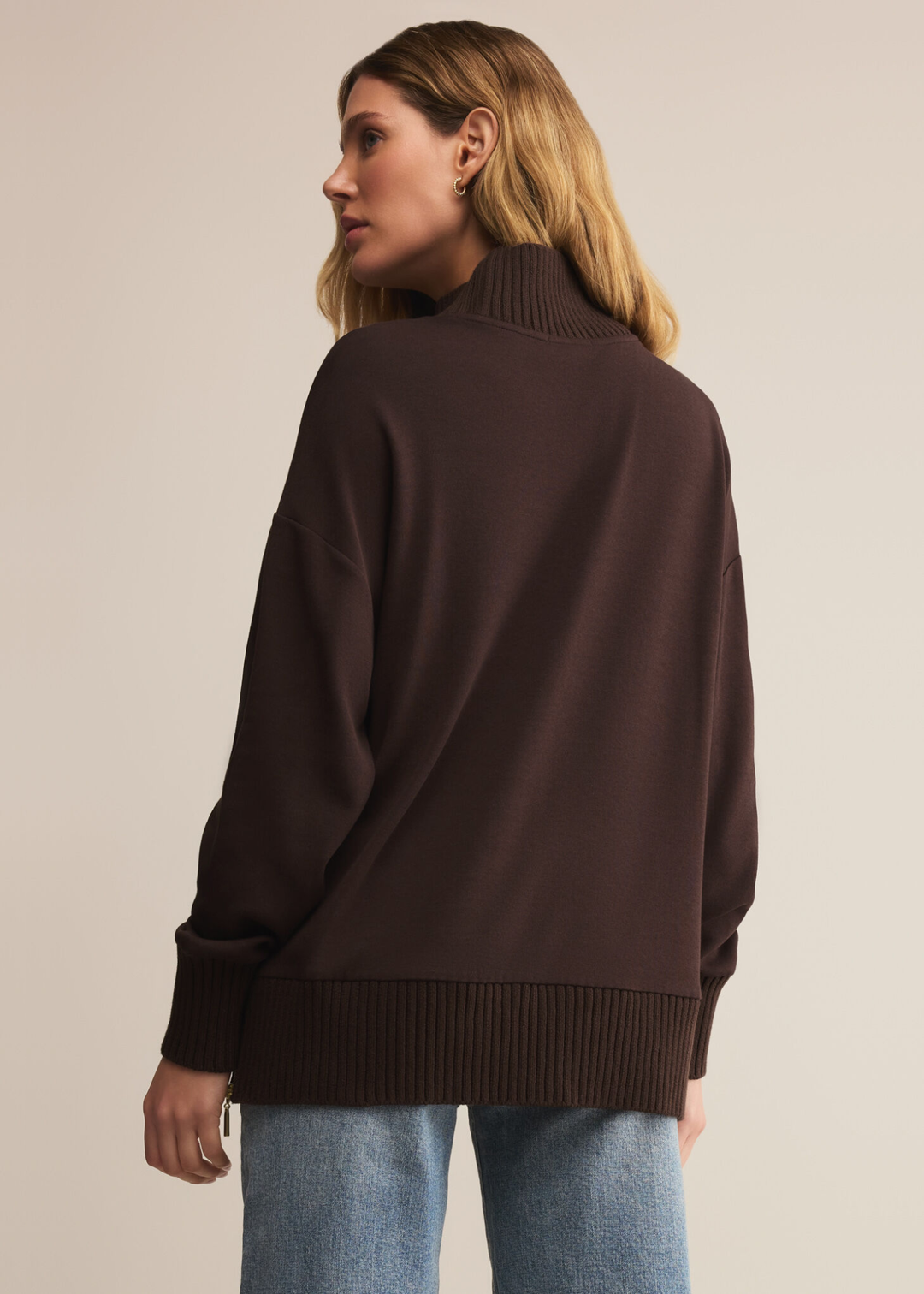 Woman turned around wearing a brown sweater with a mock neckline and jeans standing in front of a tan background