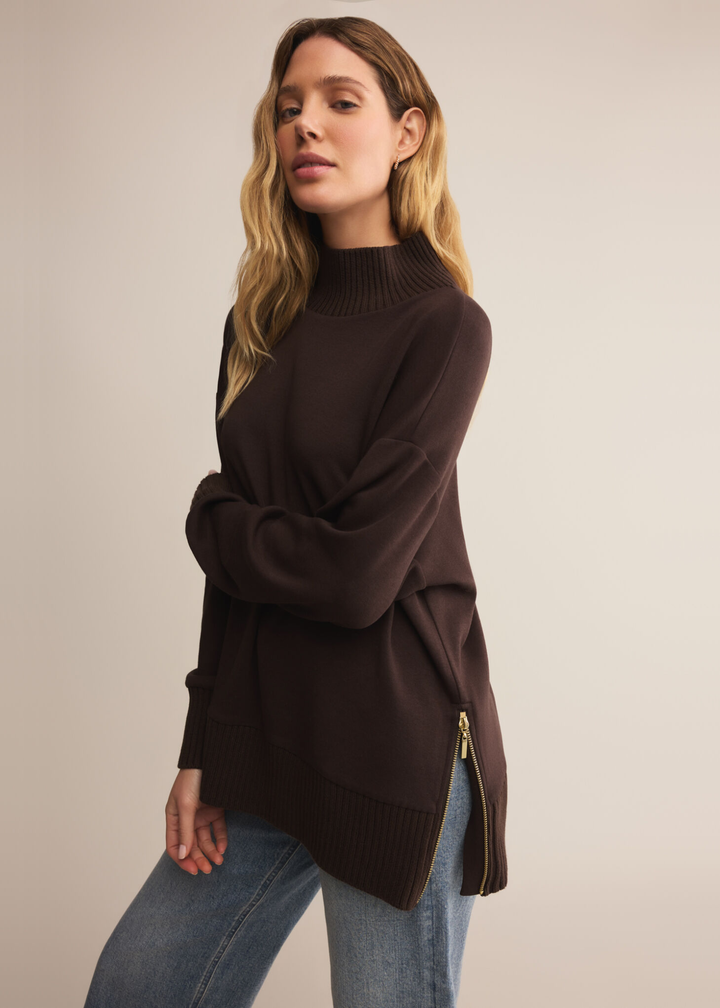 Woman wearing a brown sweater with a mock neckline and jeans standing in front of a tan background