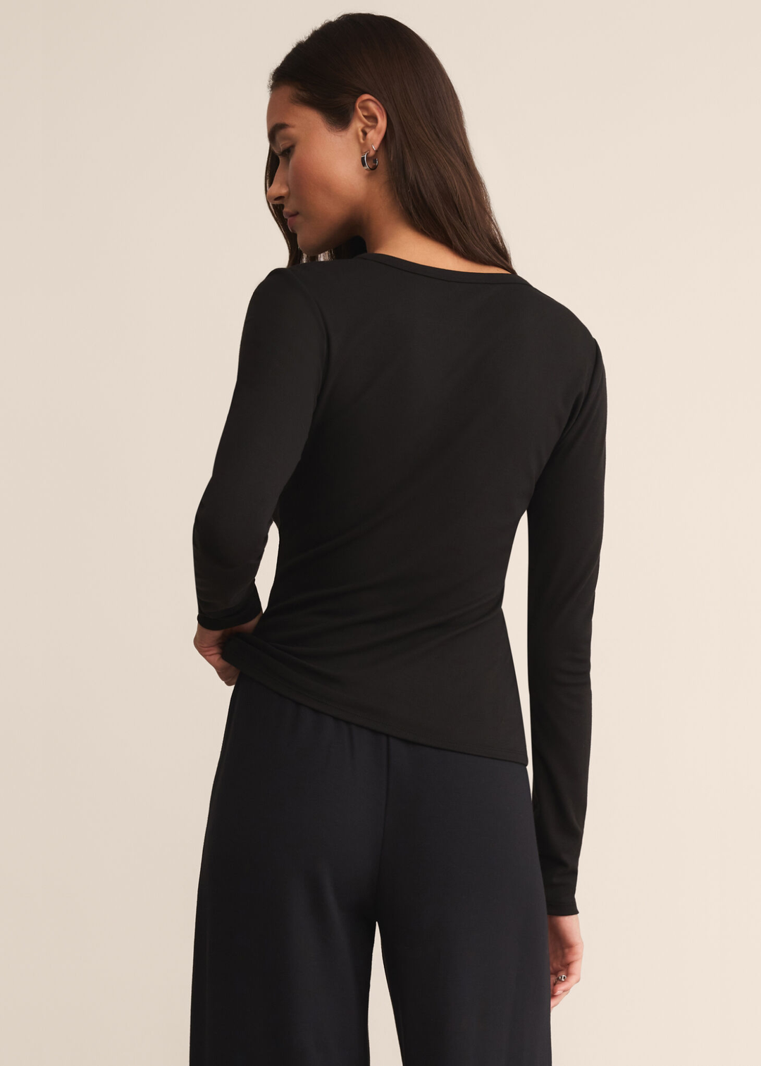 Woman turned around wearing a long sleeve black crew neck shirt with black pants and standing in front of a white background