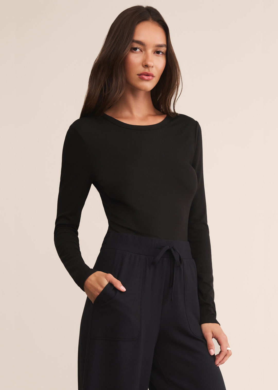 Woman with hand in pocket wearing a long sleeve black crew neck shirt with black pants and standing in front of a white background