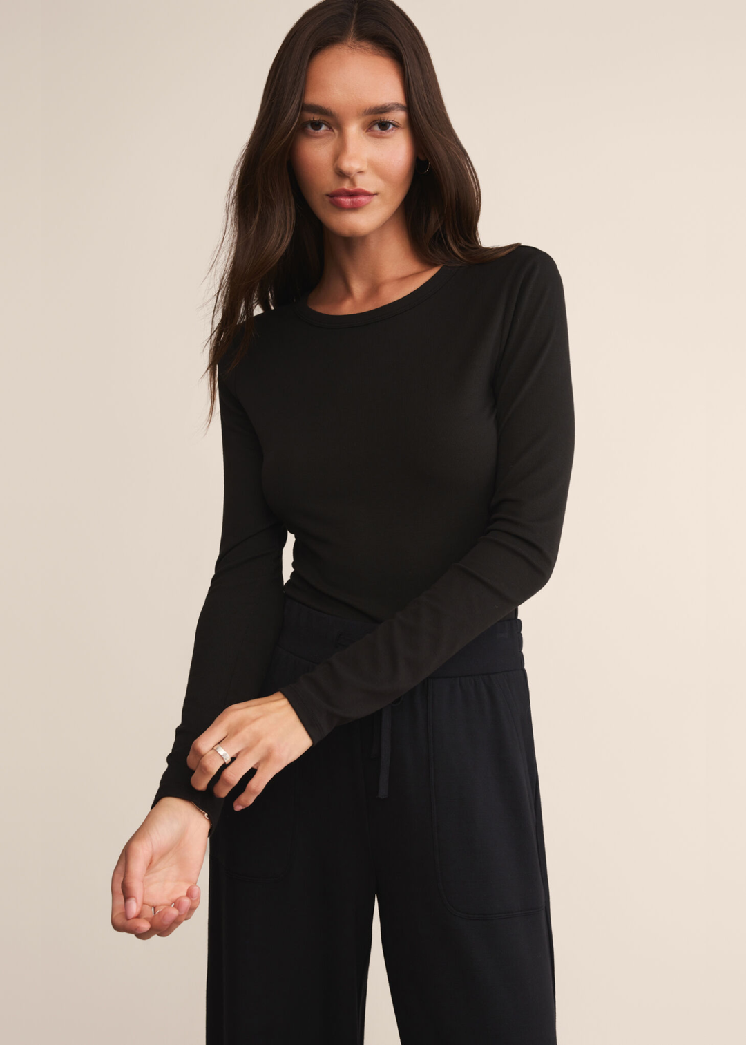Woman wearing a long sleeve black crew neck shirt and touching the sleeve with black pants and standing in front of a white background