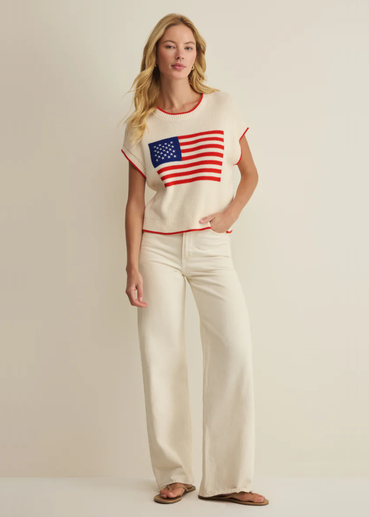Woman wearing a cream sleeveless sweater with an American flag graphic and white jeans