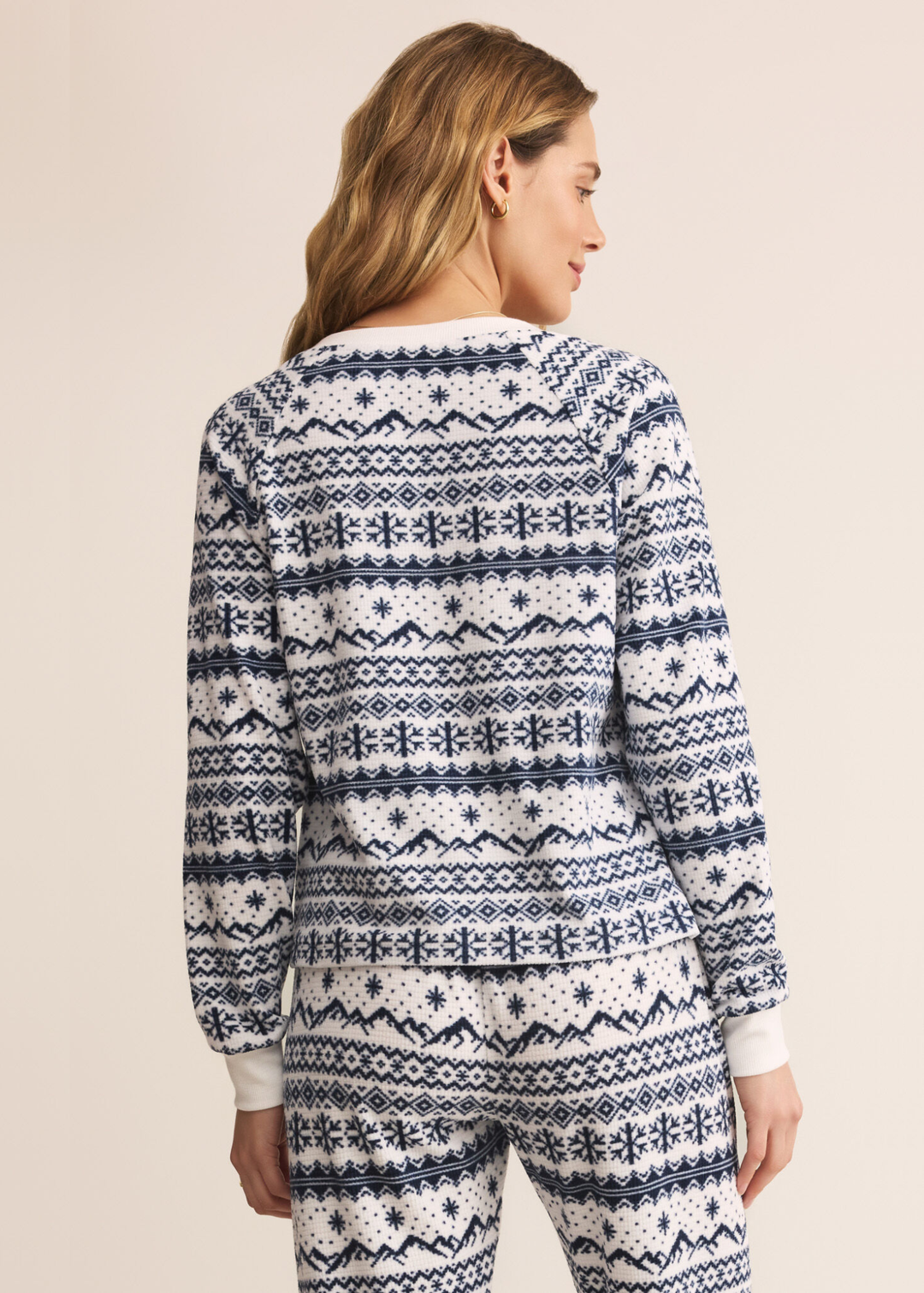 Woman turned around wearing a blue and white fairisle patterned v-neck pajama top with matching pants in front of a tan background