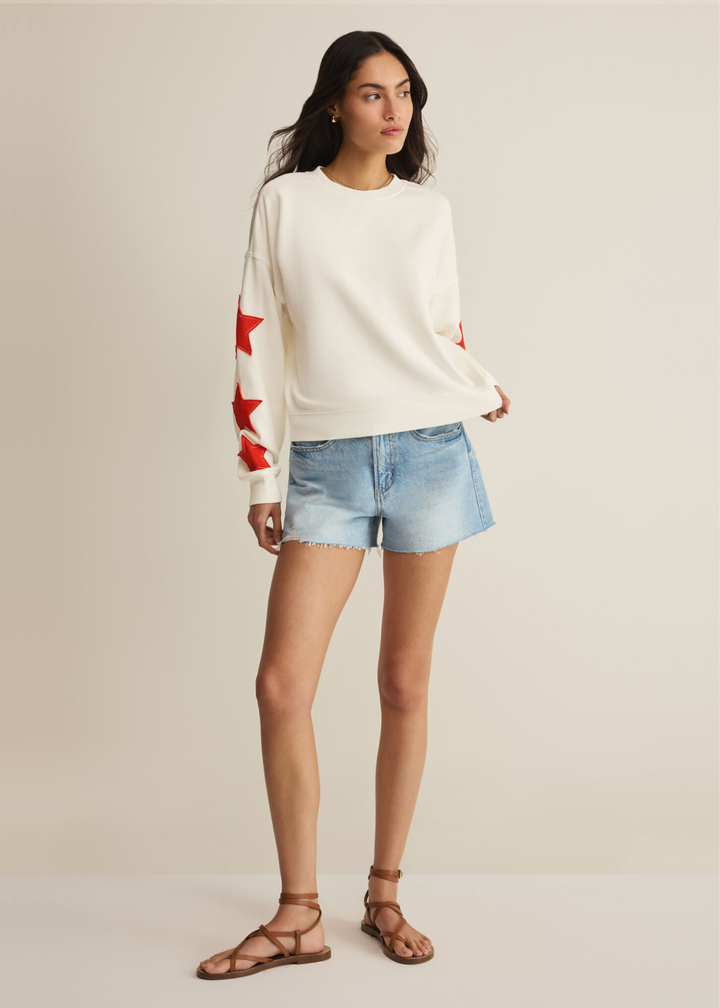 Woman wearing a cream sweatshirt with red stars stitched on the sleeves with denim shorts
