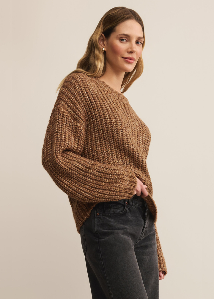 Woman turned to the side with hand in pocket wearing a brown sweater with gold flecks with black jeans standing in front of a white background