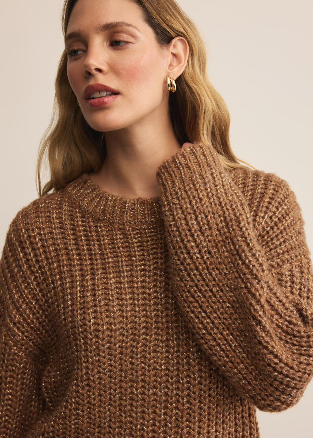 Woman wearing a brown sweater with gold flecks holding the neckline standing in front of a white background