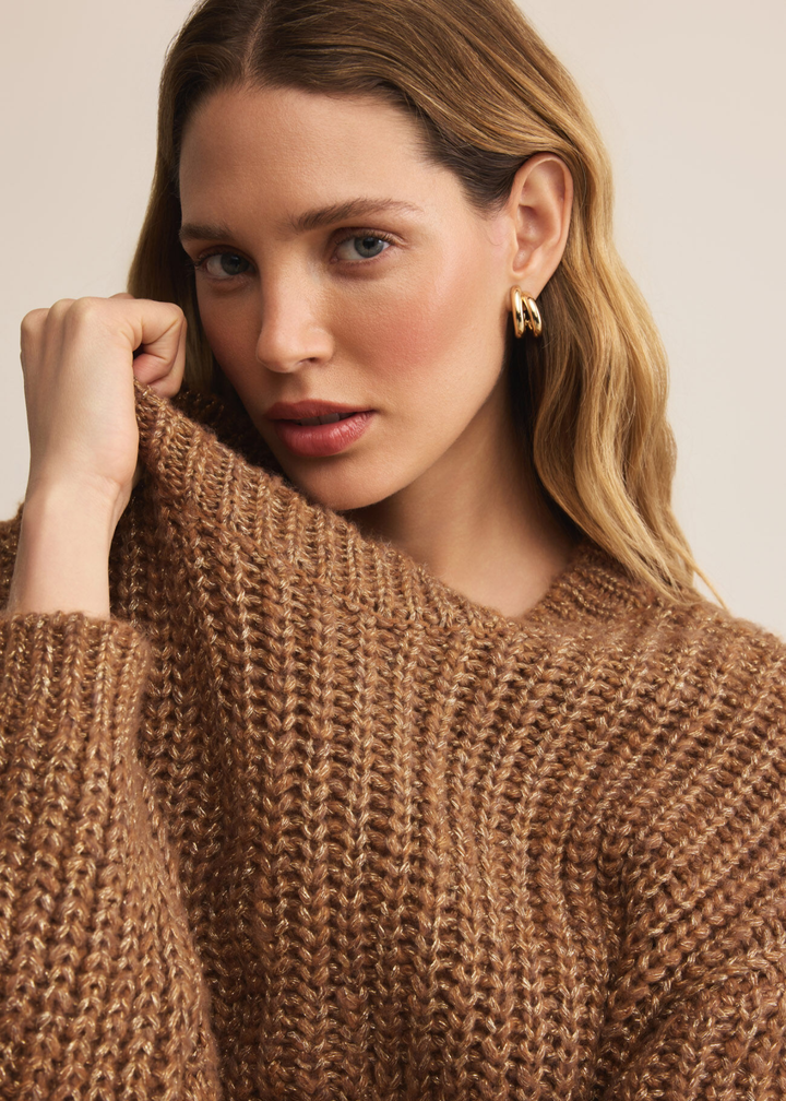 Woman wearing a brown sweater with gold flecks holding the neckline standing in front of a white background