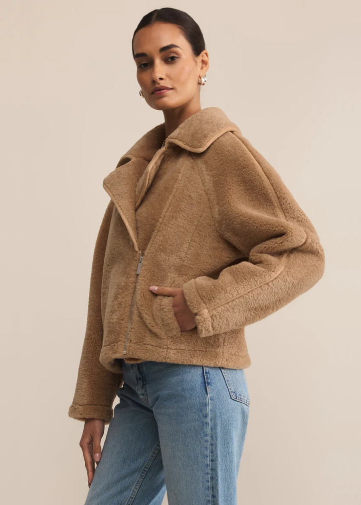 Woman turned to the side wearing a brown sherpa jacket and jeans standing in front of a tan background