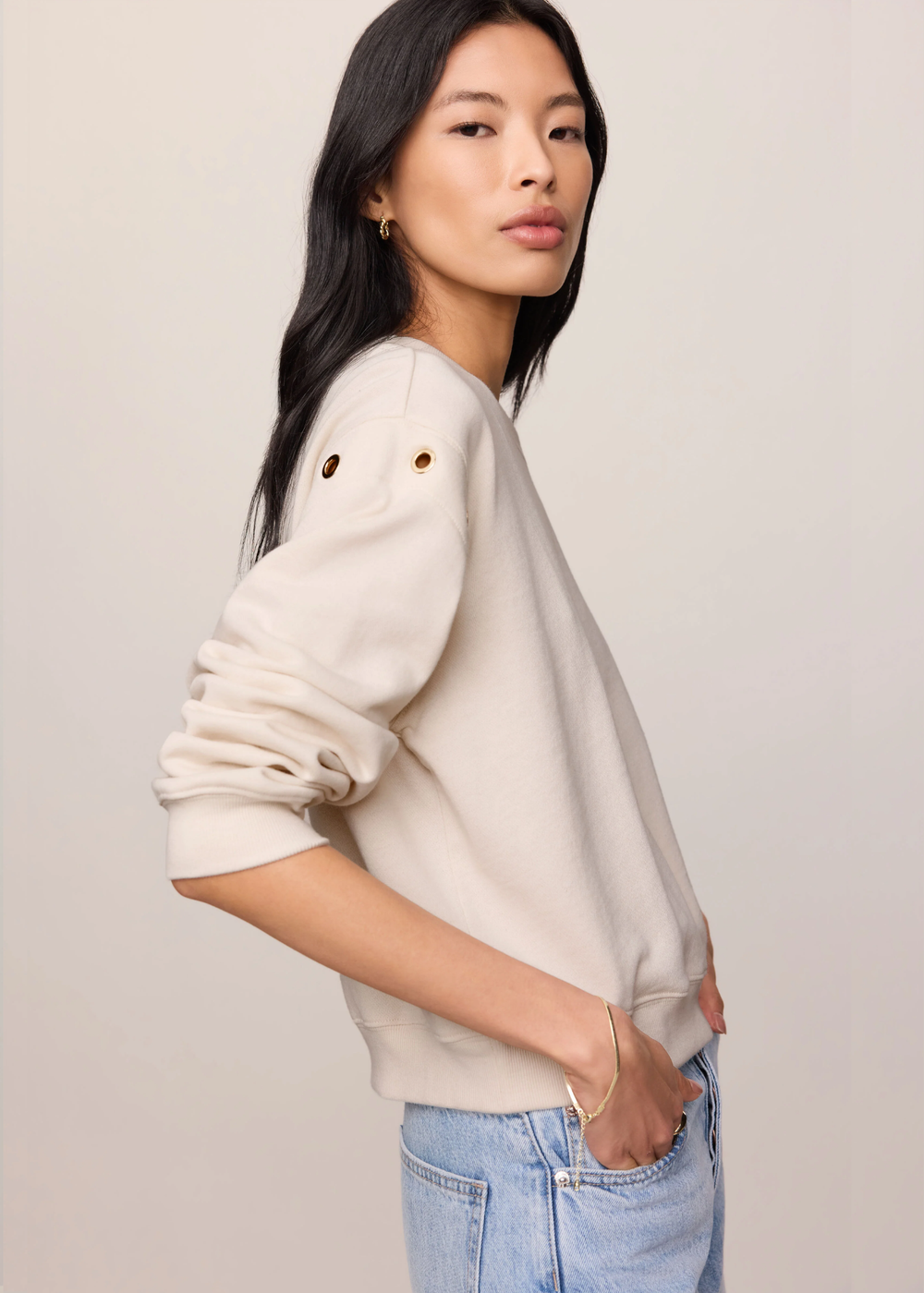A woman wearing a cream-colored crewneck sweatshirt with decorative eyelet details on the shoulders, paired with light blue jeans, photographed against a solid light beige background.
