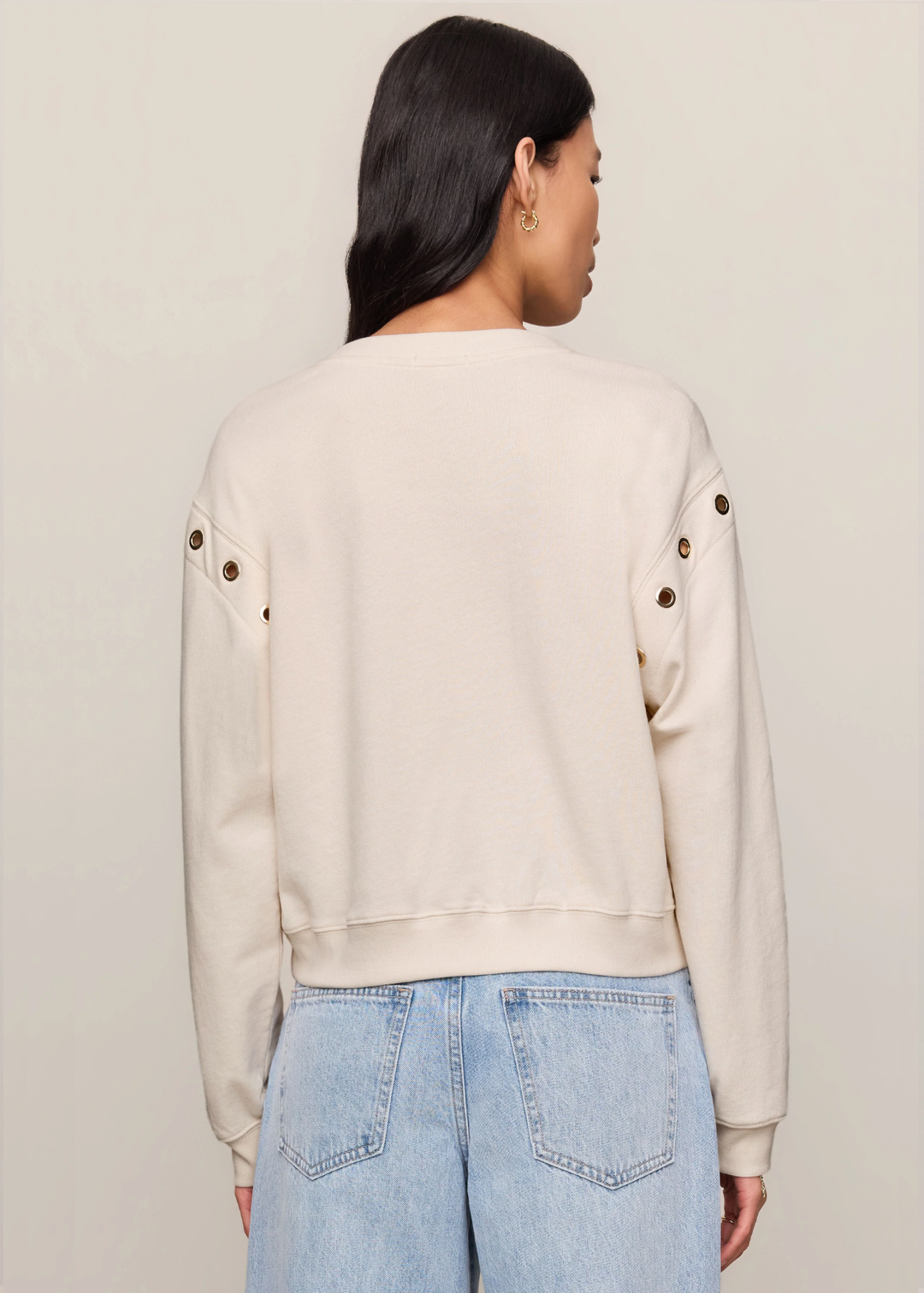 A woman shown from the back wearing a light beige crewneck sweatshirt with decorative gold-tone buttons on the sleeves, paired with light blue jeans, against a plain light beige background.