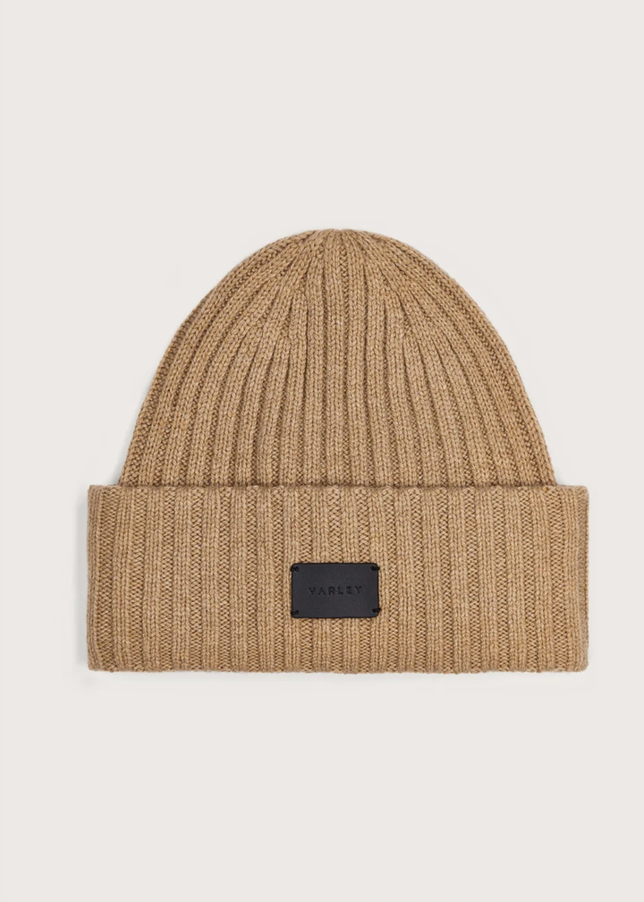 A beige ribbed knit beanie hat with a folded cuff and a small black rectangular brand patch on the front, shown against a light gray background.