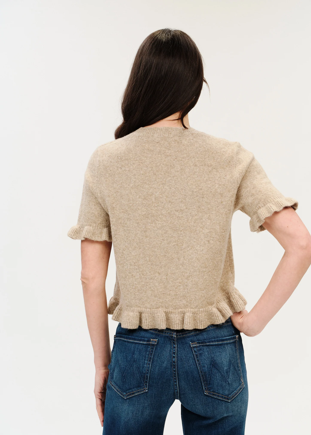 A woman viewed from the back wearing a beige short-sleeve knit top with a ruffled hem and cuffs, paired with blue jeans, against a plain light background.