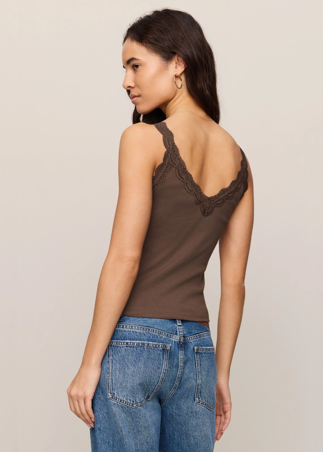 A woman shown from the back wearing a brown sleeveless camisole top with lace trim along the open back, paired with blue jeans, against a plain light beige studio background.