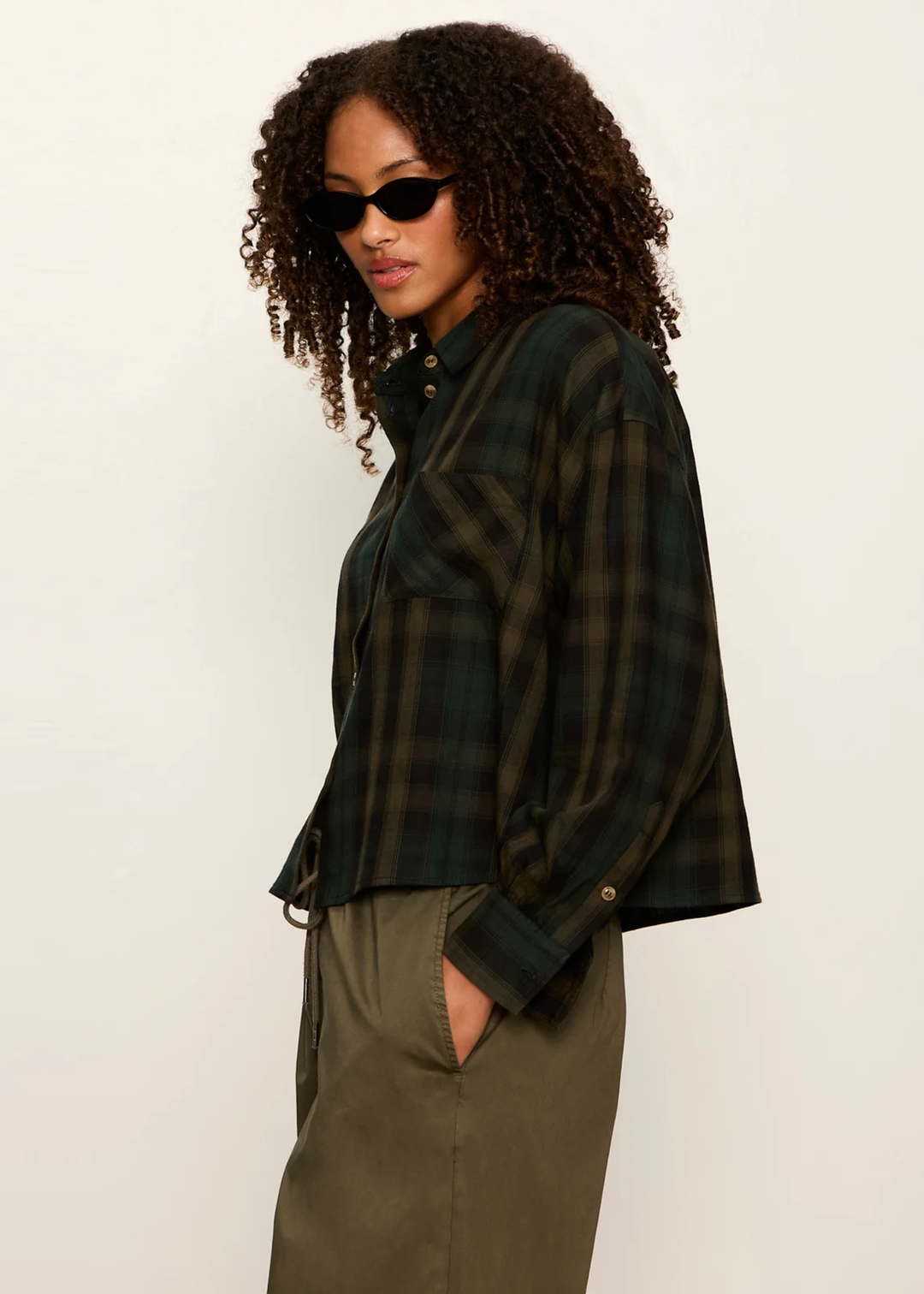 A woman with curly hair wearing a dark green and black plaid button-up shirt and olive green pants, photographed against a plain light background.